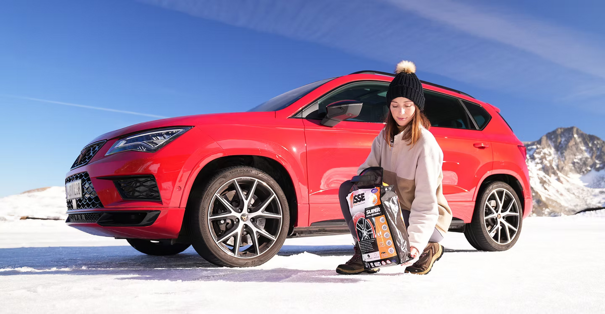 Mujer agachada junto a un coche rojo sobre la nieve sosteniendo una bolsa de fundas textiles para nieve ISSE Chains Super.